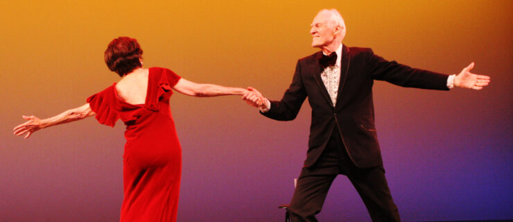 Stuart Hodes, in a black tuxedo, lunges sideways, holding the hand of a woman in a red dress as she leans away from him, balanced on one foot as if ending a turn. They are onstage with an orange backdrop behind them.