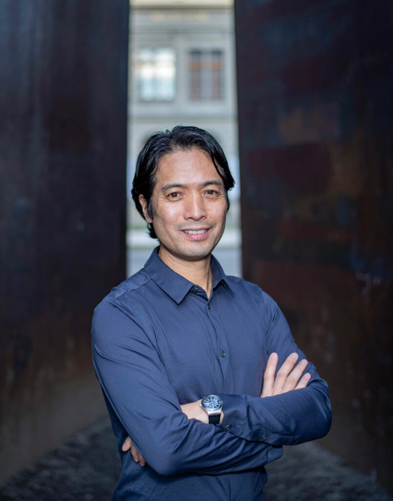 Kinsun Chan, a middle-aged man of East Asian descent, smiles at the camera with his arms crossed. His black hair is mostly pushed back from his face. He wears a medium blue button down shirt.
