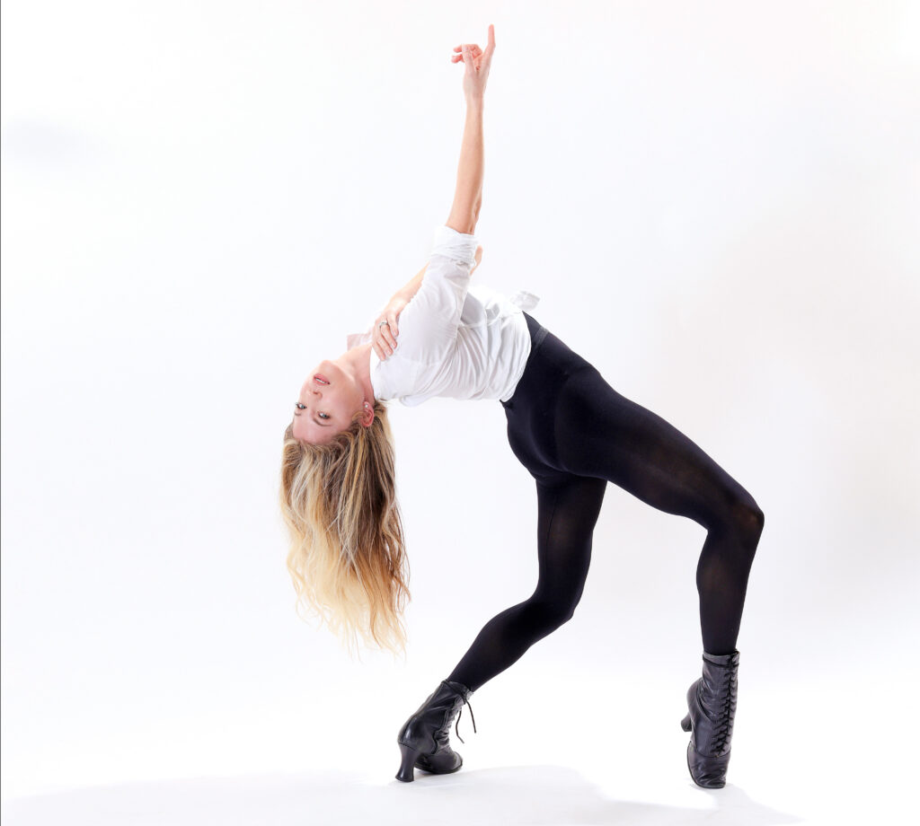 Ashley Blair Fitzgerald, a young white woman, poses against a white backdrop. She gazes at the camera upside down, long legs in plié as she arches into a backbend with one arm reaching to the sky. Her blonde hair nearly reaches her back heel. She wears a white button-down tied at the waist, sleeves rolled up, over a skintight black unitard and black lace-up jazz boots.