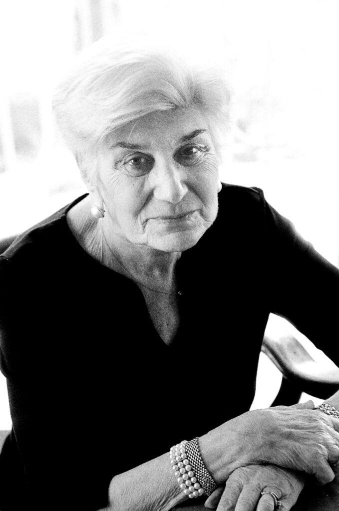 an elderly woman smiling at the camera , wearing a dark dress and pearl jewelry 