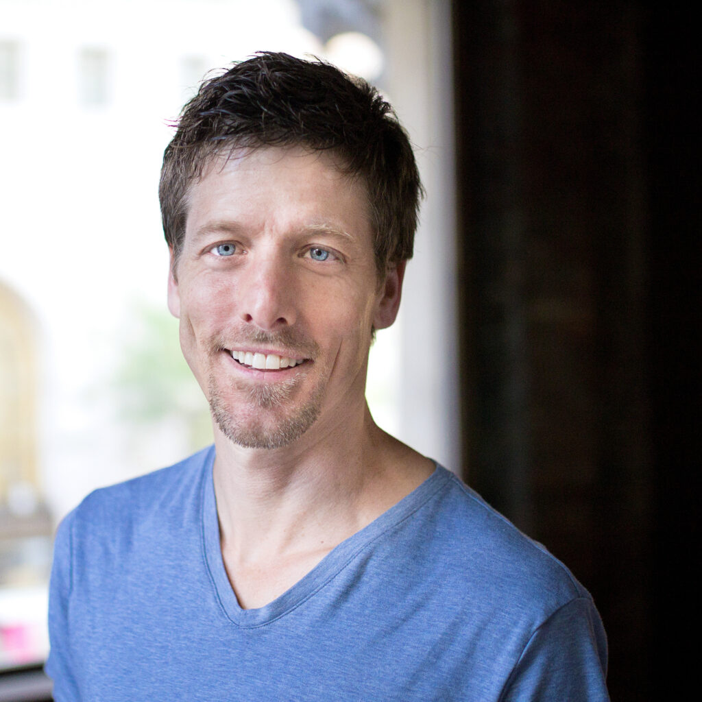 a male with brown hair wearing a blue t shirt smiling at the camera