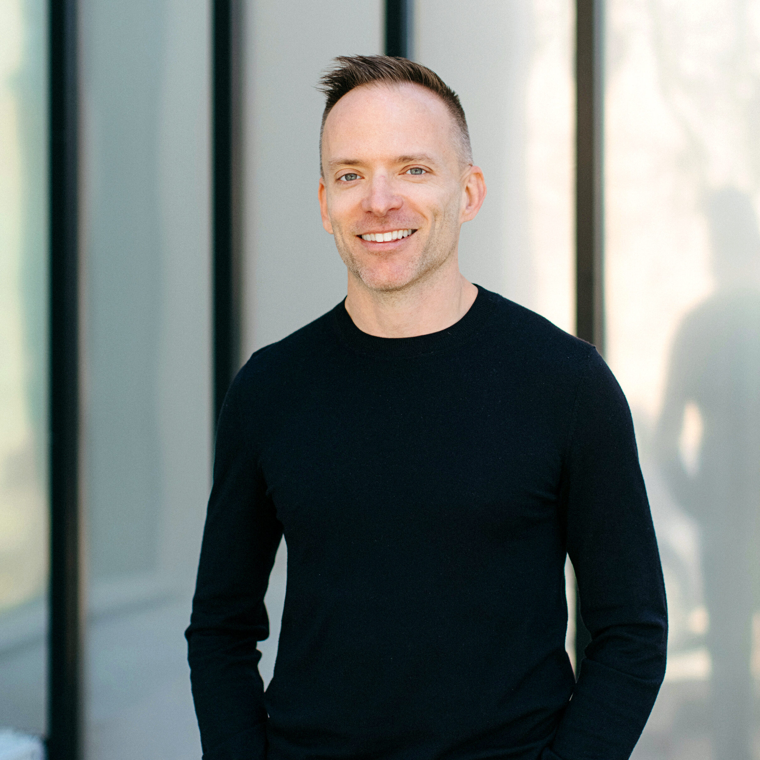 Ryan Donovan, a white man with brown hair, stands smiling with his hand in his pockets. He wears a black long sleeve shirt and jeans. He is outside, in front of a cream colored building with vertical black accents. 