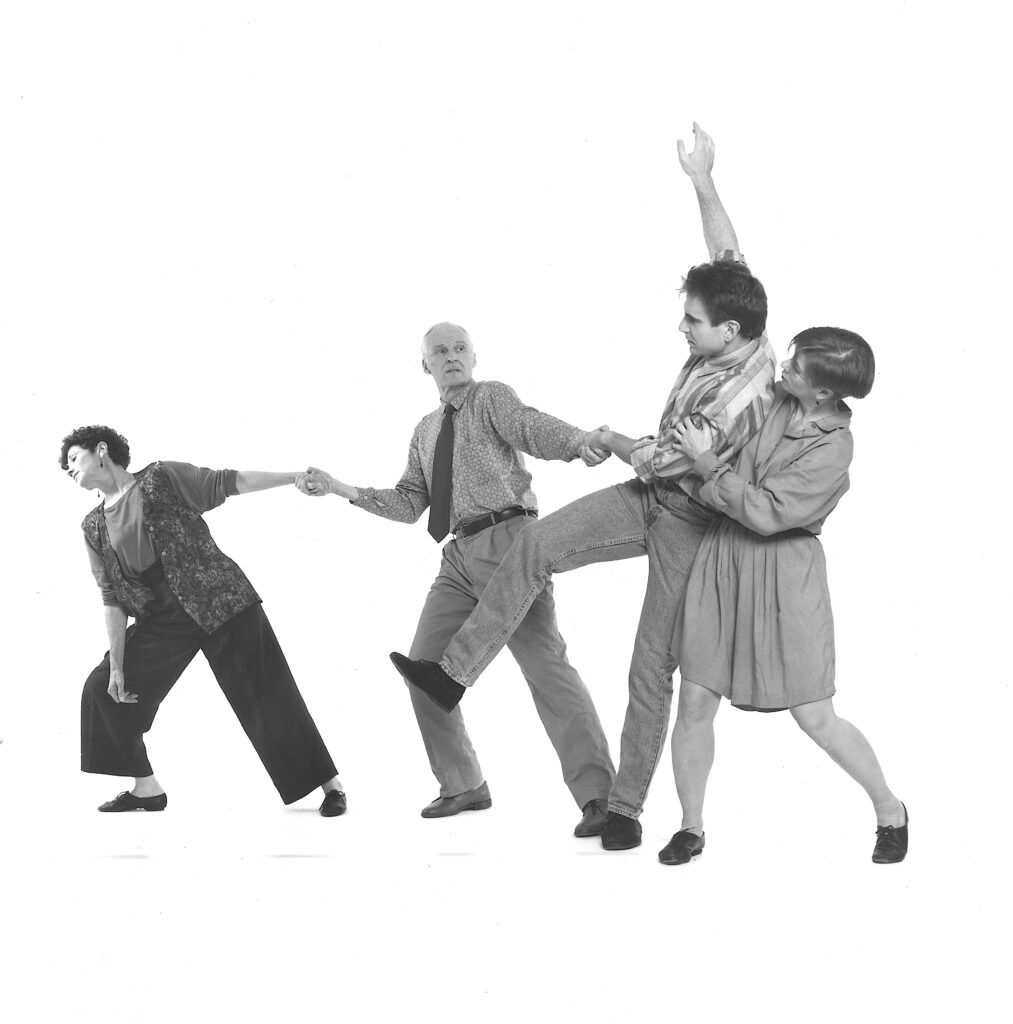 A black and white images of four dancers connected in a horizontal line, the left two holding hands and leaning toward the left. The dancer on the right supports the second from right as he lifts one arm and leg and leans into her.