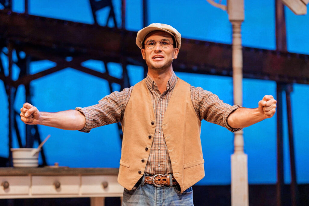 male performer wearing brown vest, plaid shirt, and newsboy hat, arms outstretched 