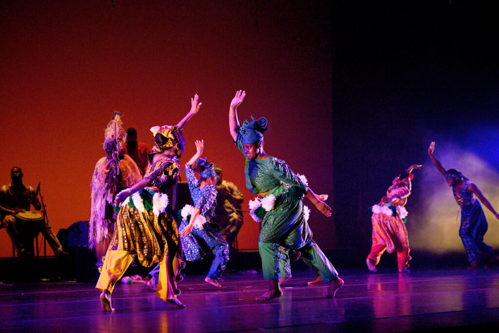 Two dancers downstage lunge toward each other, upstage arms raising high overhead in a swing. Other brightly costumed dancers do the same in pairs upstage.