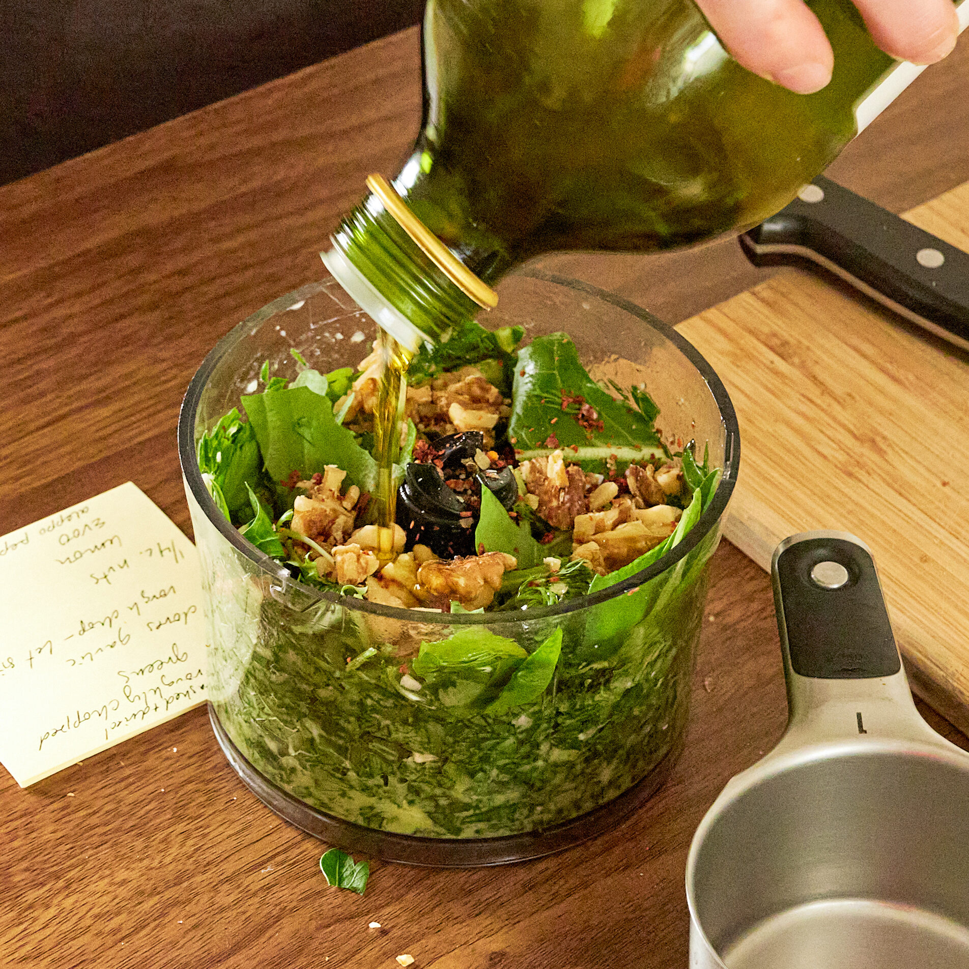 a blender with green, nuts, and herbs in it with olive oil being poured over top