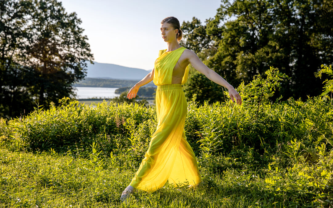 female dancer in a long yellow dress standing in the grass with water behind her