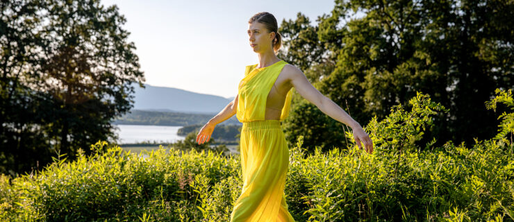 female dancer in a long yellow dress standing in the grass with water behind her