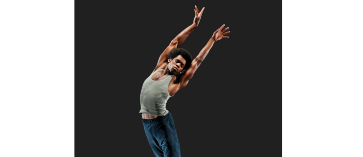 a male dancer wearing a tank top, pants, and jazz sneakers jumping in the air