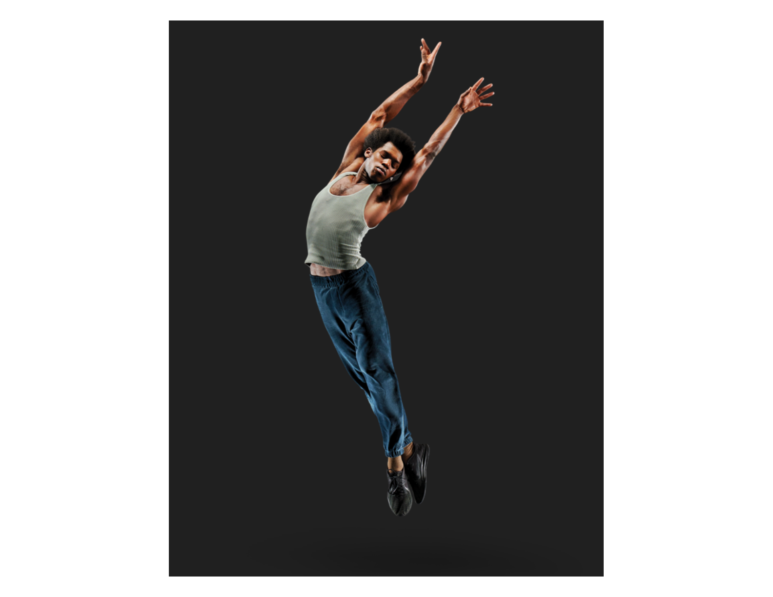 a male dancer wearing a tank top, pants, and jazz sneakers jumping in the air