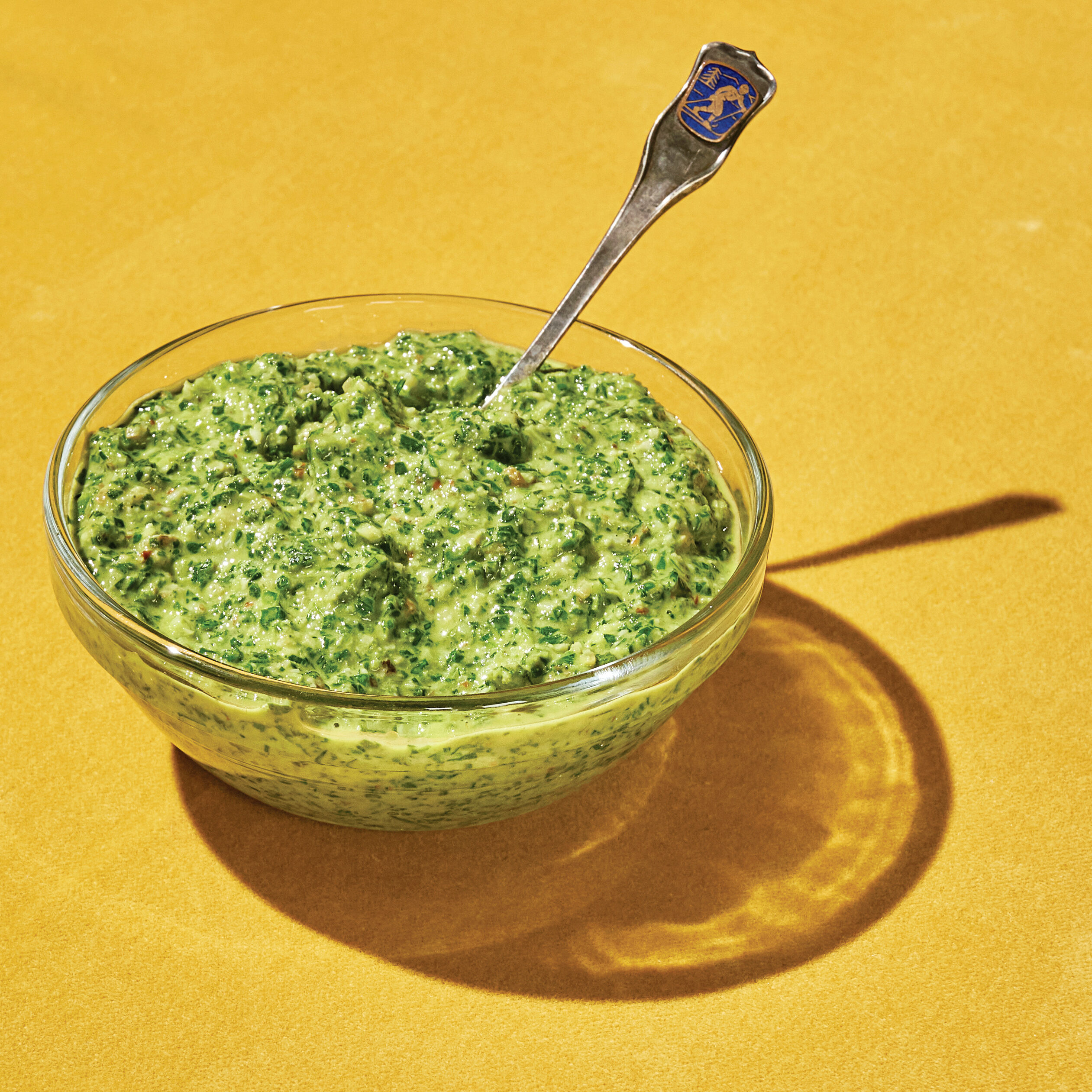 green pesto inside a clear bowl with a silver spoon against a yellow background