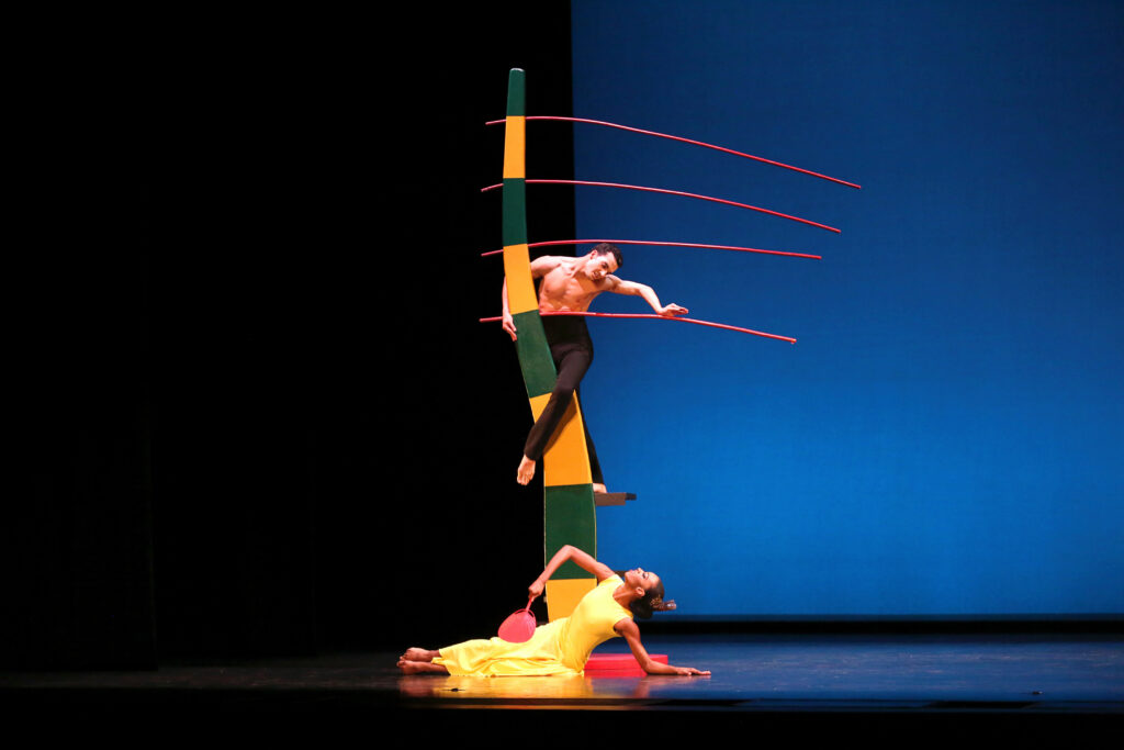 A male dancer climbs a whimsical, curving tower of thick green and gold stripes, four orange-red rods extending straight to the side. He rests the heel of a cupped hand on one of these rods as he gazes down at a dancer in yellow seated at the base of the tower. She holds a red fan as she reclines on one elbow, the other elbow jabbing upward.