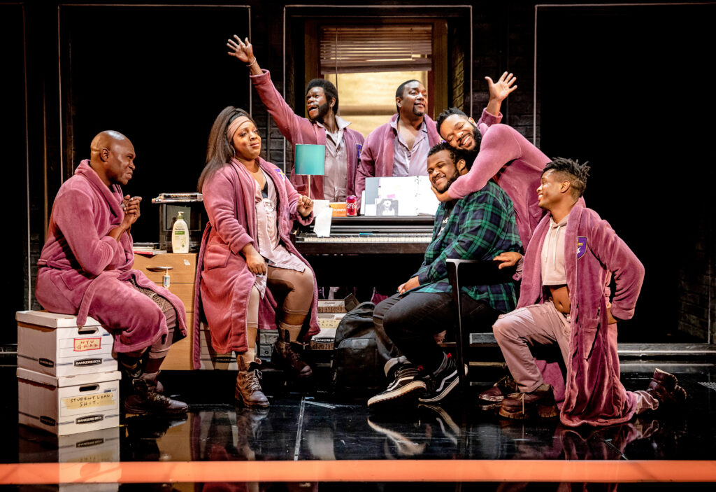 A Strange Loop's main character, Usher, sits at a keyboard in the suggestion of a cluttered, cramped New York City apartment, surrounded by six pink-clad ensemble members representing his thoughts. One hugs him from behind with a big smile, while two behind the desk extend jazz hands on opposite diagonals. The other three are perched around the space, either smiling or looking at him in entreaty.