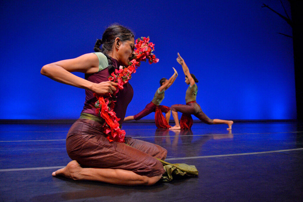 three dancers on stage, one holding a long rope of lei of flowers