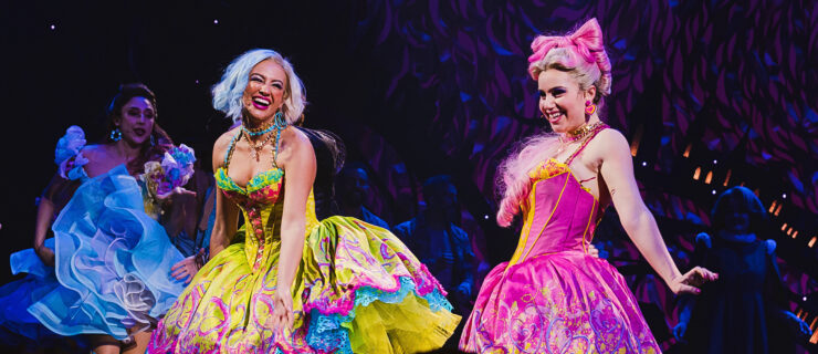 A performance photo from the musical Bad Cinderella. A performer in an over the top hot pink dress and an oversized bow to match poses to make the fluffy skirt of her dress swirl, with a bright smile, while another in chartreuse laughs, looking over her shoulder at her.