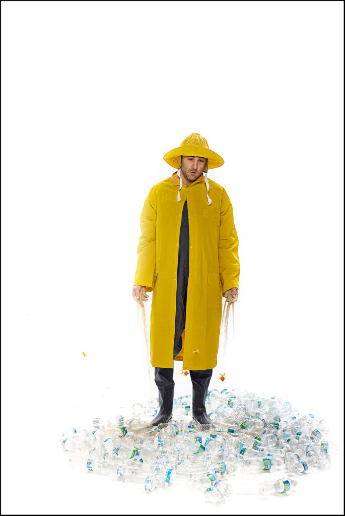 A man in a bright yellow raincoat and matching hat despondently holds nets filled with empty plastic water bottles.