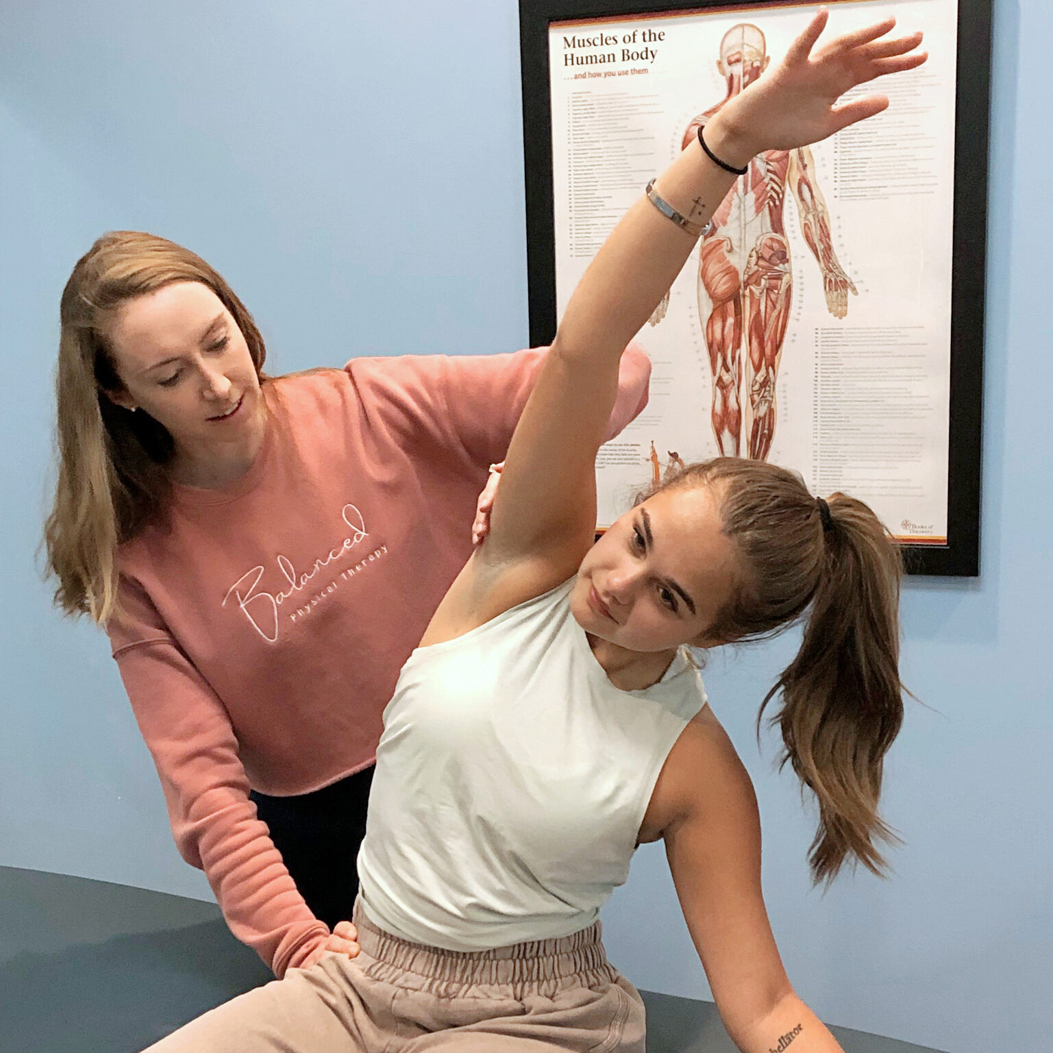a female physical therapist wearing a pink sweatshirt helping a female dancer stretch laterally on an examination table