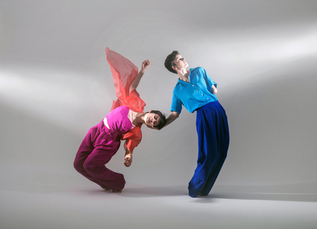 Two dancers pose against a grey backdrop. Caitlin Hicks, in reds and pinks, hinges toward the floor, listing toward her left side. Julie Crothers catches her head with one hand as she faces away, her body mirroring Caitlin's arc.