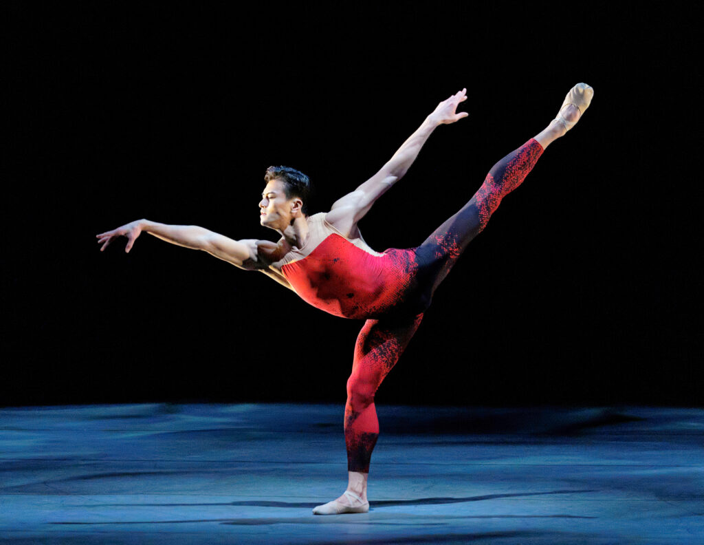 Chun Wai Chan stretches into an exaggerated fondu arabesque, torso and front arm reaching forward against his high back leg. He wears a fitted unitard that evokes lava with its red and black pattern.