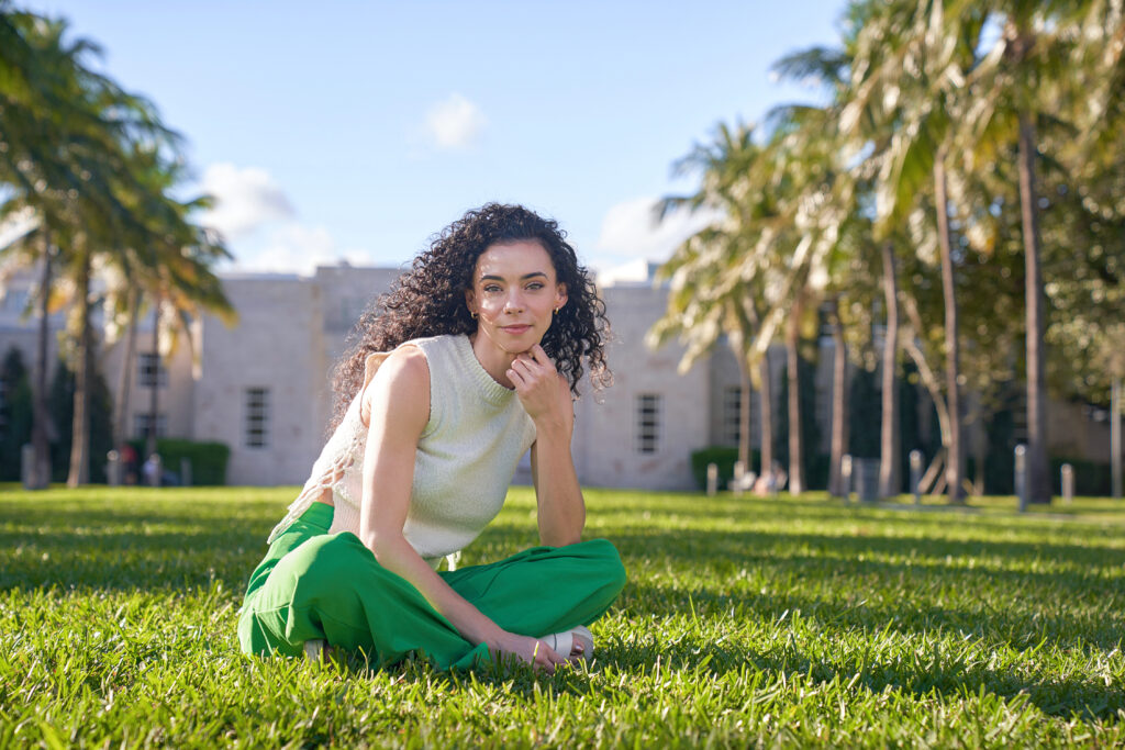 a female wearing a white tank top and green pants sitting cross-legged in the grass