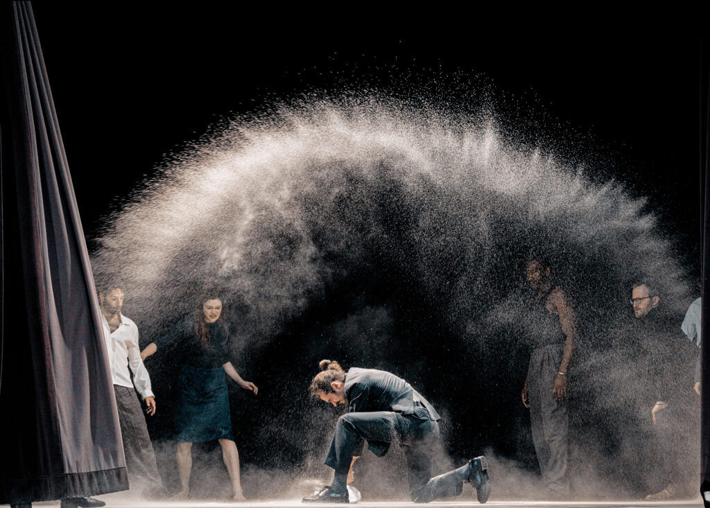 A massive spray of grey powder flies into the air as a male dancer throws a bag to the ground, kneeling over it. Other dancers on the periphery watch or flinch away from the motion.