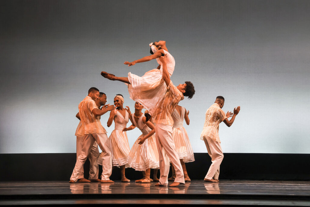 A female dancer is lifted from below her shoulders, head arcing back toward the ceiling and both legs raised in attitude back. A half-dozen other dancers are visible upstage, keeping up a beat as they clap and dance with each other. All wear white dresses or shirts and trousers that evoke the mid-twentieth century. The women's pointe shoes are dyed to match their skin tones.