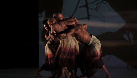 A small group of Black dancers, all shirtless and wearing calf-length skirts that fade from white to deep red, cluster together. They face inward, carefully cradling necks or backs with arms and hands, a moment of care in the midst of motion. The backdrop gives the impression of trees in silhouette.