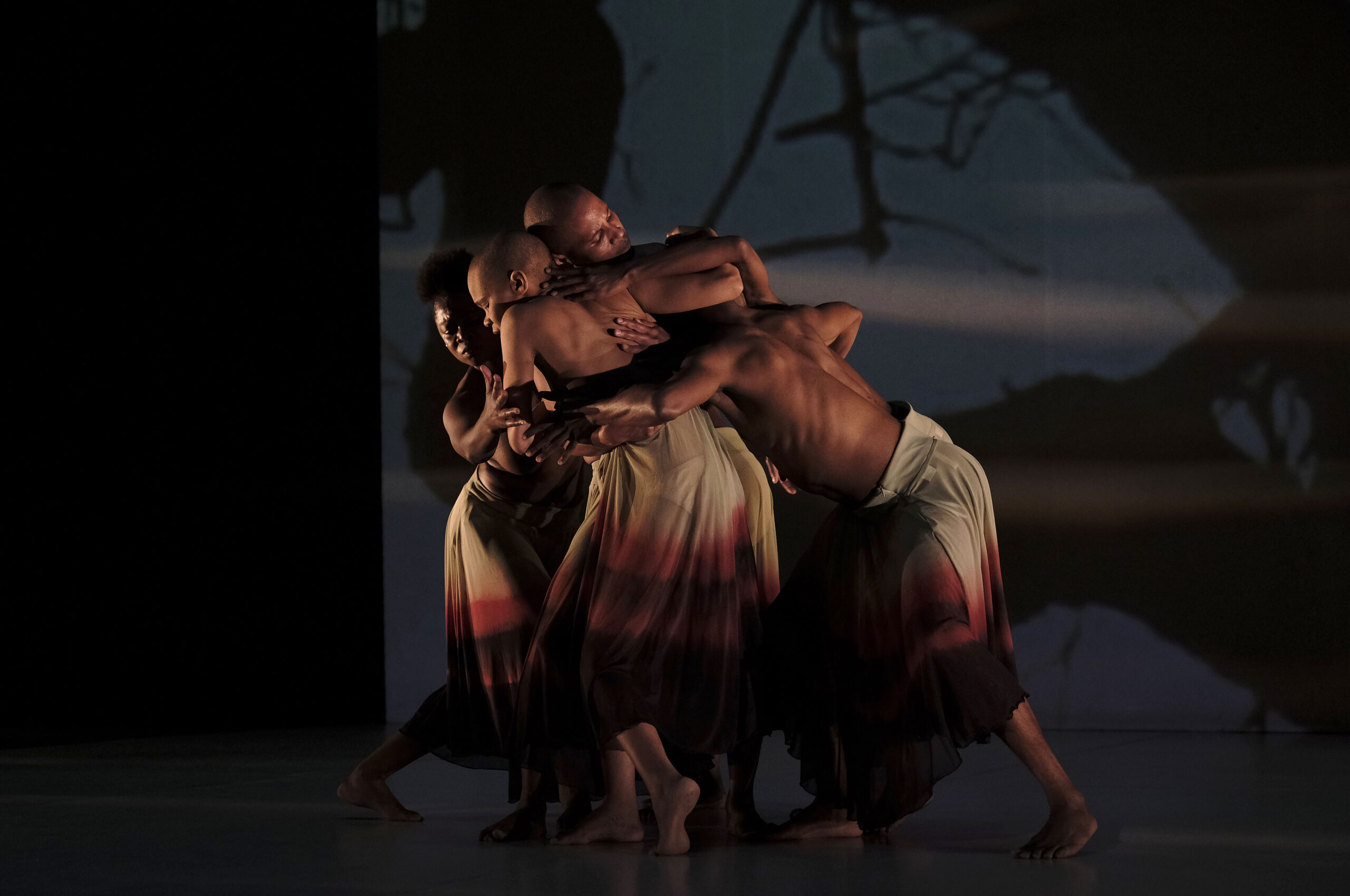 A small group of Black dancers, all shirtless and wearing calf-length skirts that fade from white to deep red, cluster together. They face inward, carefully cradling necks or backs with arms and hands, a moment of care in the midst of motion. The backdrop gives the impression of trees in silhouette.