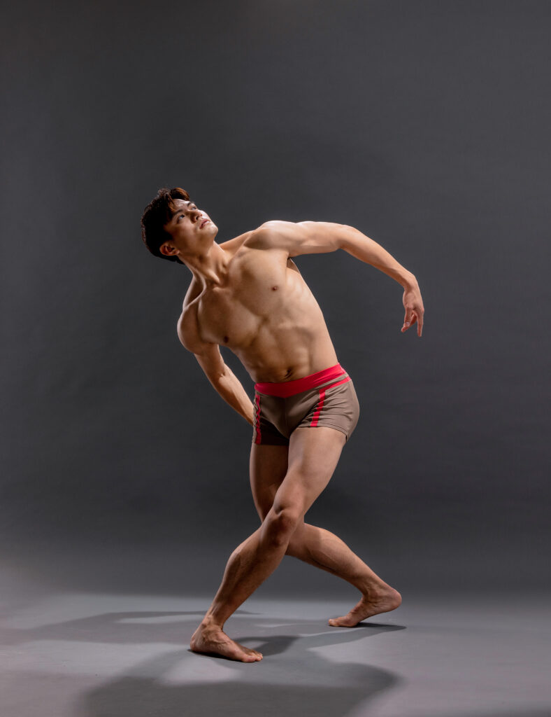 Chun Wai Chan, a muscular Chinese man in his early 30s, poses against a grey backdrop. He pliés in fourth position, back heel popped. His arms curve to form a circle just behind his torso as he leans to his right, gazing up over his upraised left shoulder. He is barefoot and bare-chested, and wears red and grey patterned shorts.