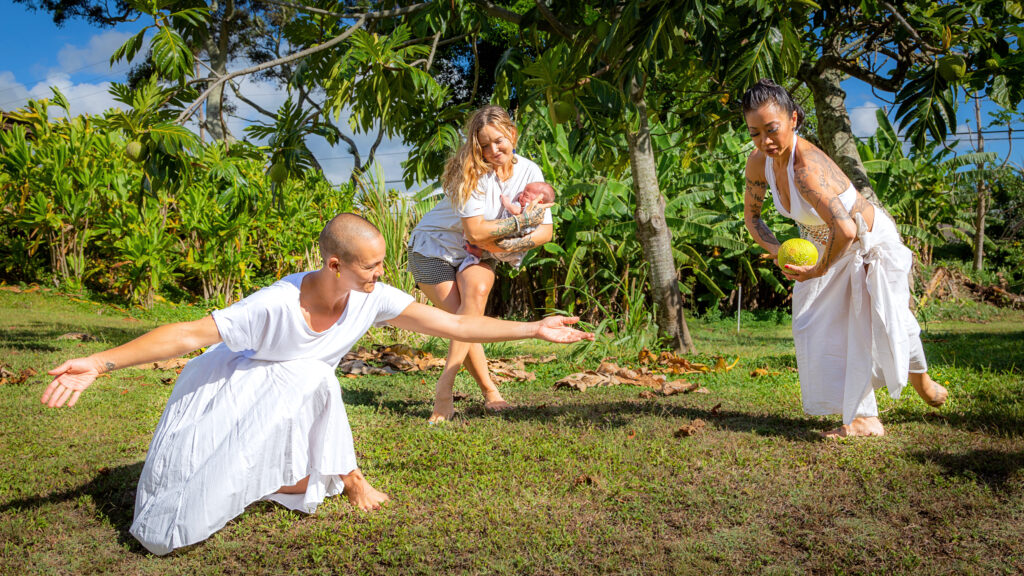 three dancers performing in the grass all wearing white, one is holding a small baby