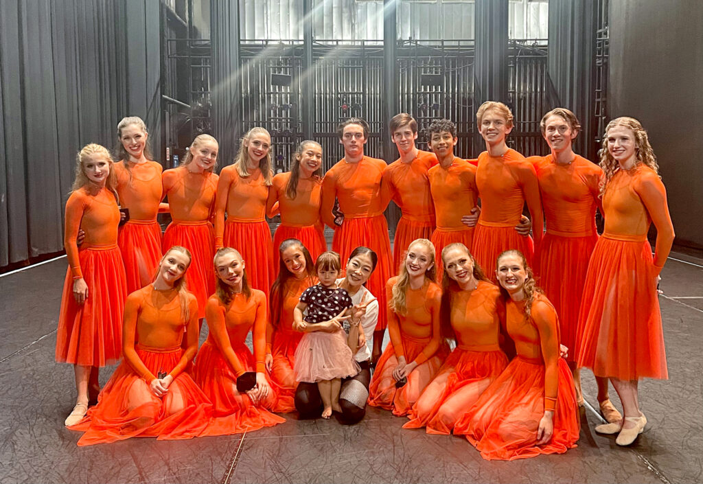 a woman and small girl smiling for a photo with a large group of dancers wearing orange costumes