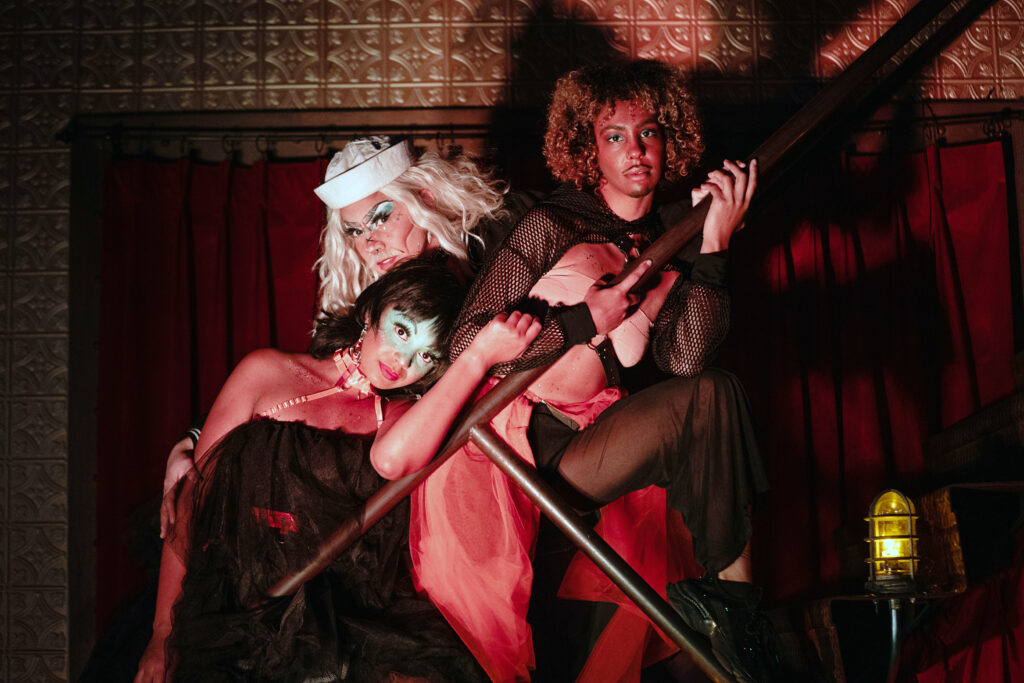 Three performers pose on a metal stair in a richly decorated, red-lit room. They hold each other as they gaze at the camera, wearing a motley assemblage of face paint, sailor caps, fishnets, and harnesses.