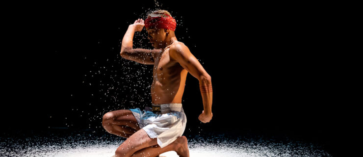 a male dancer kneeling on a bed of rice with rice falling down on him