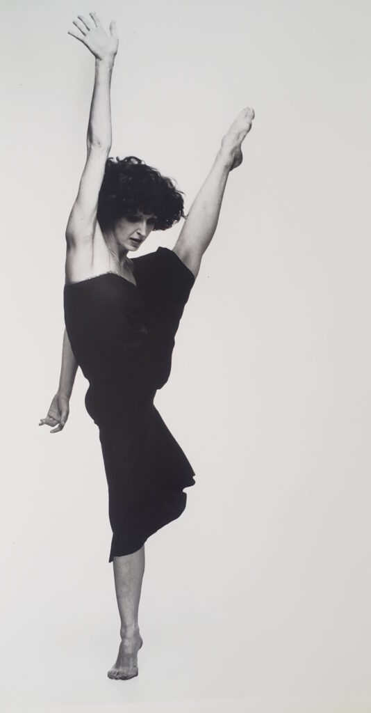 In a black and white archival image, Jennifer Muller balances on relevé, one leg extended high to the side. Her opposite arm reaches high, palm splayed and shoulder rising. Her chin ducks slightly. She wears a dark jumpsuit that ends at the knees, the fabric flowing with the motion. Her curly hair is down and flies around her head.