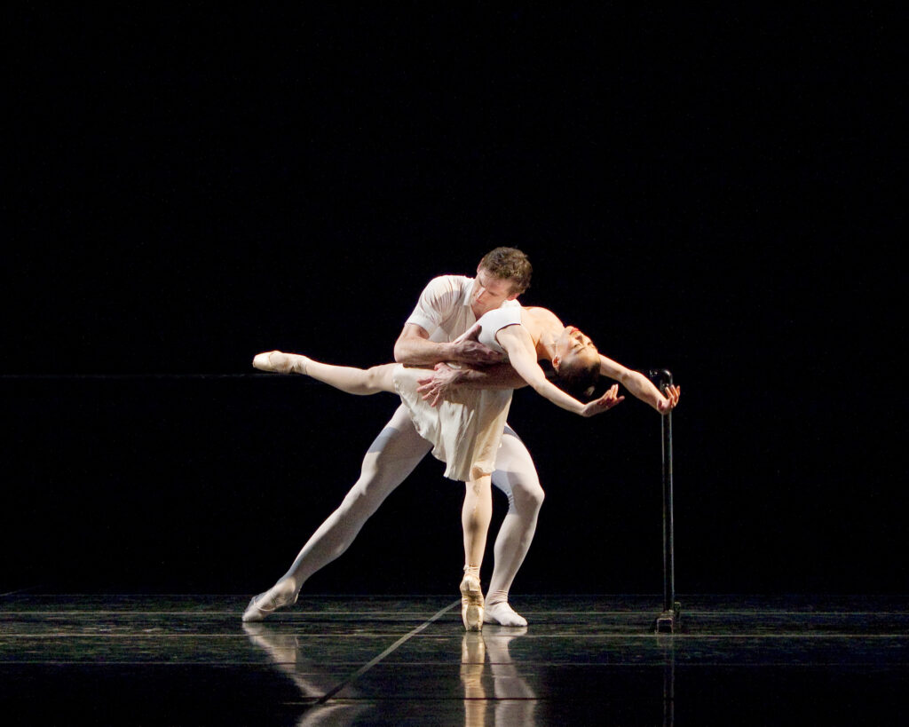 A dancer in pink tights and pointe shoes and a white skirt and leotard ensemble arches back, arms overhead, as she balances en pointe with her leg extended ninety degrees to the front. Her partner, in tights and a white tunic, lunges to support her at the waist as he tenderly touches his head to her ribcage.