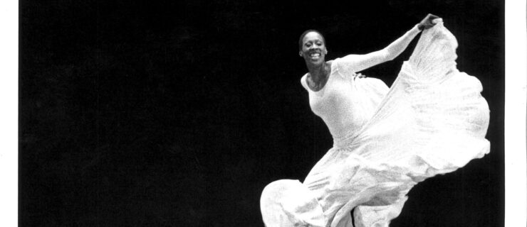 In a black and white archival image, Judith Jamison grins infectiously as she dances. She wears a long white dress, the skirt of which she lifts into a ripple with one arm as she moves lithely on the balls of her feet, knees bent.