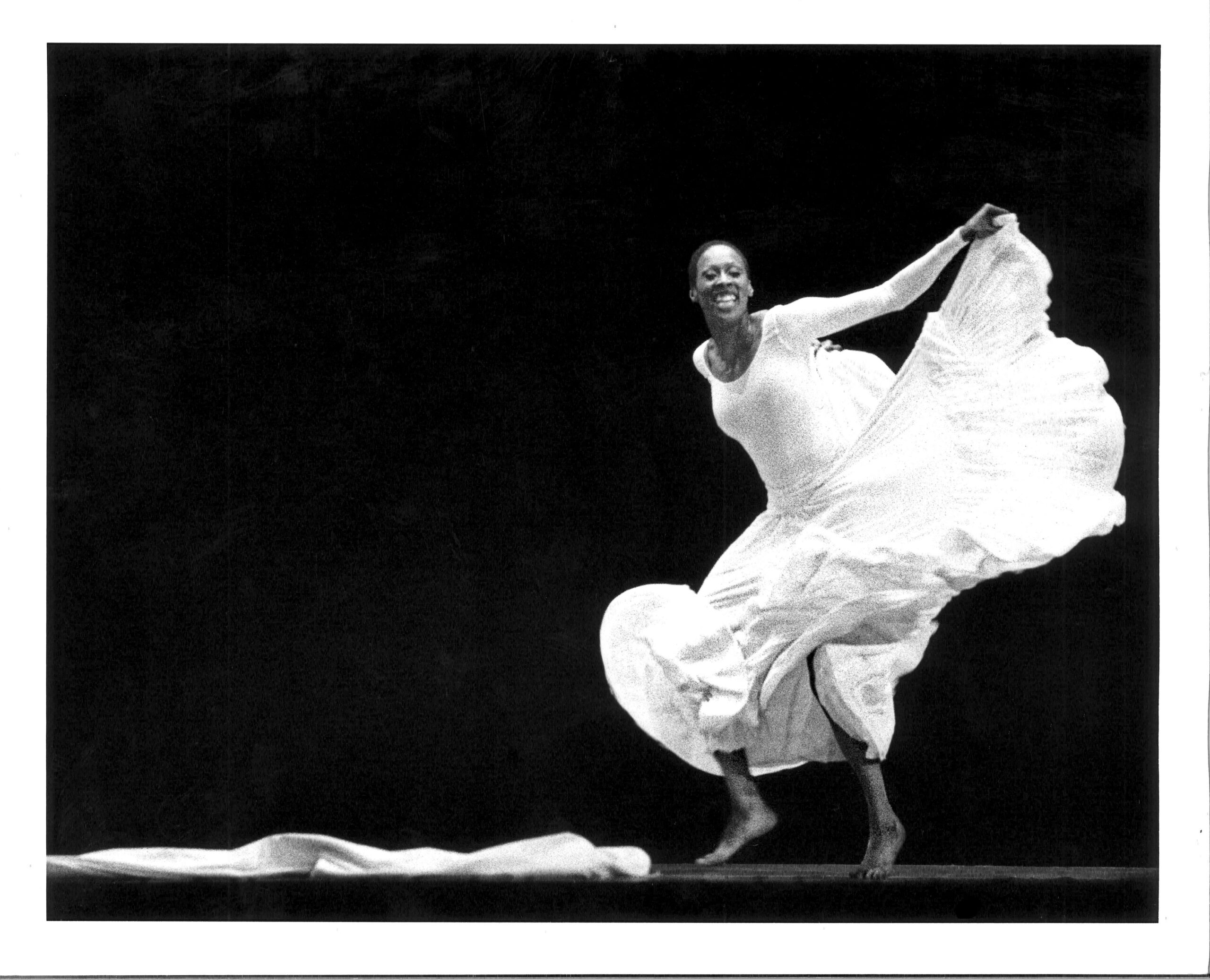 In a black and white archival image, Judith Jamison grins infectiously as she dances. She wears a long white dress, the skirt of which she lifts into a ripple with one arm as she moves lithely on the balls of her feet, knees bent.