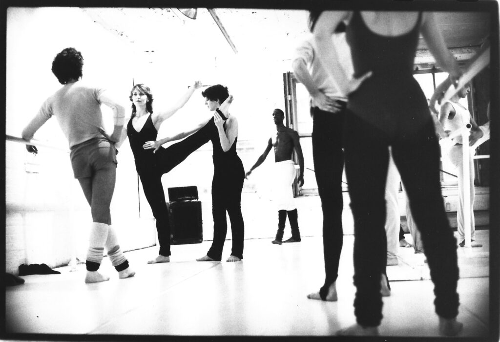 In a black and white archival image, Jennifer Muller stands beside a dancer at barre with their leg extended side. Muller holds the leg with hand at the ankle while using the other to touch the dancer's hip. Other figures in practice clothes look on, hands on hips.
