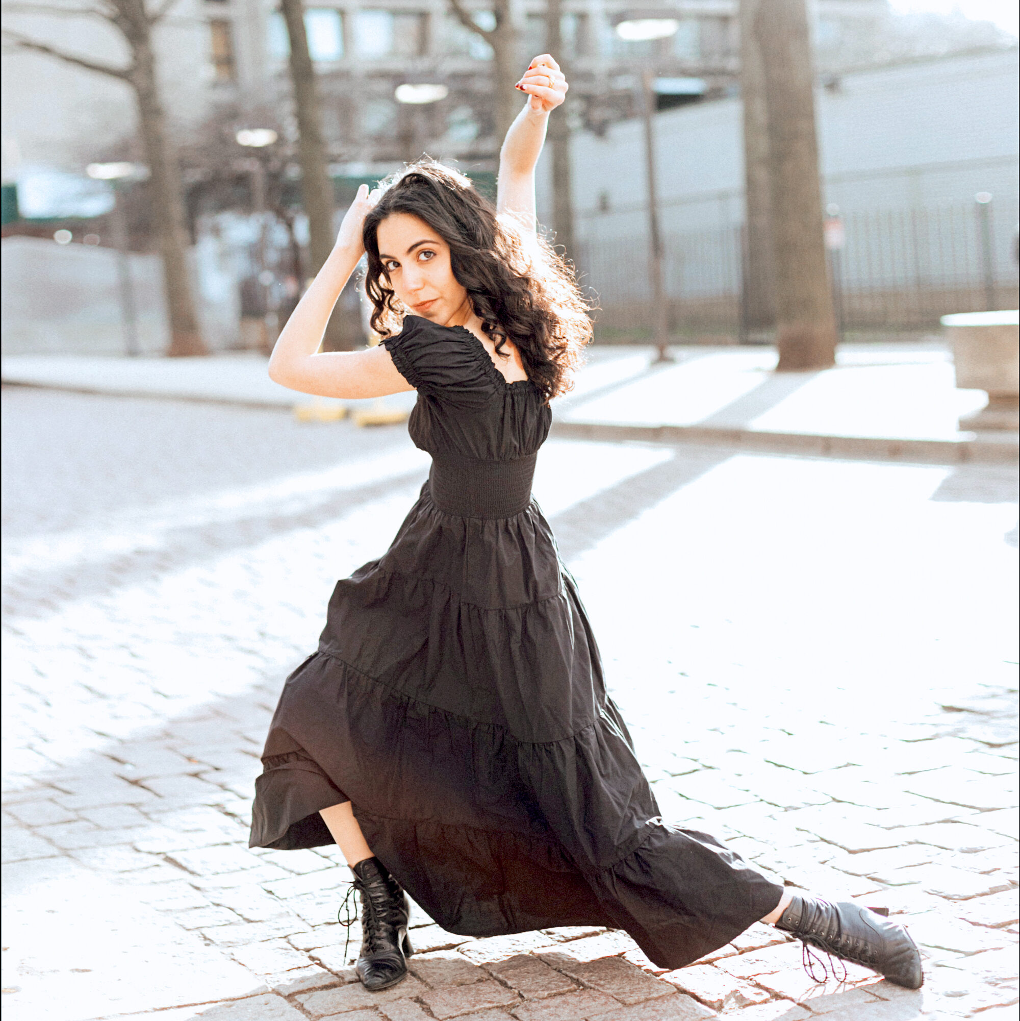 a female dancer with dark curly hair wearing a black dress and dancing in the street 
