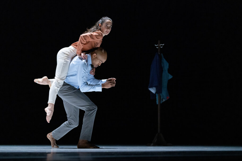 A dancer hoists herself onto the back of her partner as he curves forward with bent knees. Both wear business casual attire; a couple of jackets are visible on a coat rack that is in the shadows upstage.