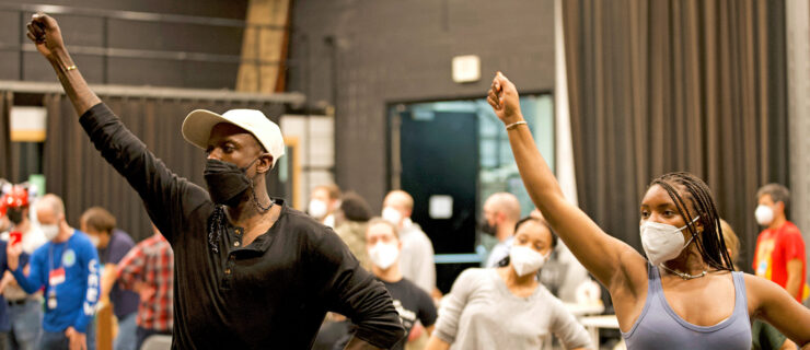 Raja Feather Kelly demonstrates in a rehearsal room, one hand upraised as though it holds a lit torch, same-side hip hitching upward. He wears a white cap, a black face mask, a black t-shirt, and sweats.