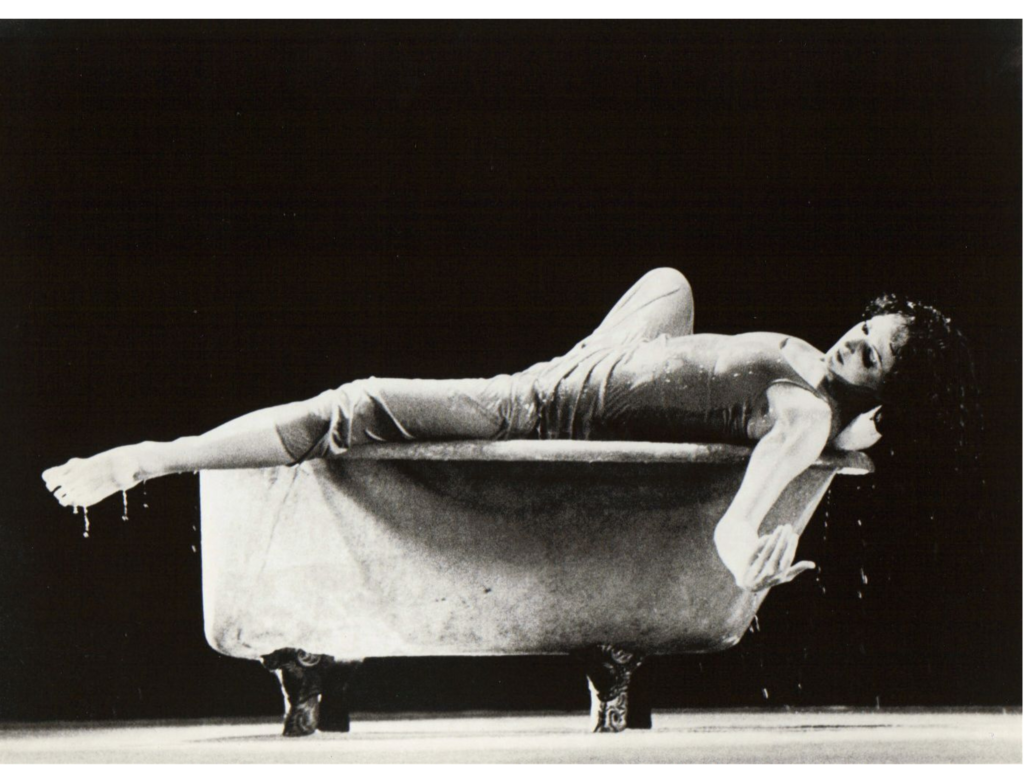 In a black and white archival image, Jennifer Muller lies atop a freestanding bathtub, balancing on the edges. Water drips from her downstage foot, extended elegantly past the front of the tub. She glances over her downstage shoulder, arms gracefully extended down and to her side, palms open.