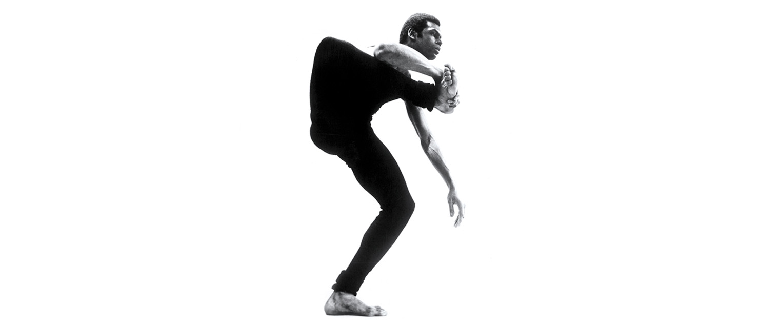 In a black and white archival photo, a young Gus Solomons jr, all long, lanky limbs, poses against a white backdrop. He balances on one leg, knee bent, as he raises the other knee up to his shoulder and grasps his flexed foot with one hand. His upper torso curves forward as he looks to the right, his free arm dangling elegantly at his side.