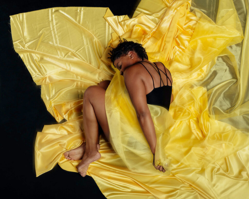 Leslie Parker lies on her side with her knees tucked up to her chest, upper arm resting long against the length of her torso. Yellow fabric is draped over her hips and underneath her. Her head is tucked under, eyes closed, as though she is asleep.
