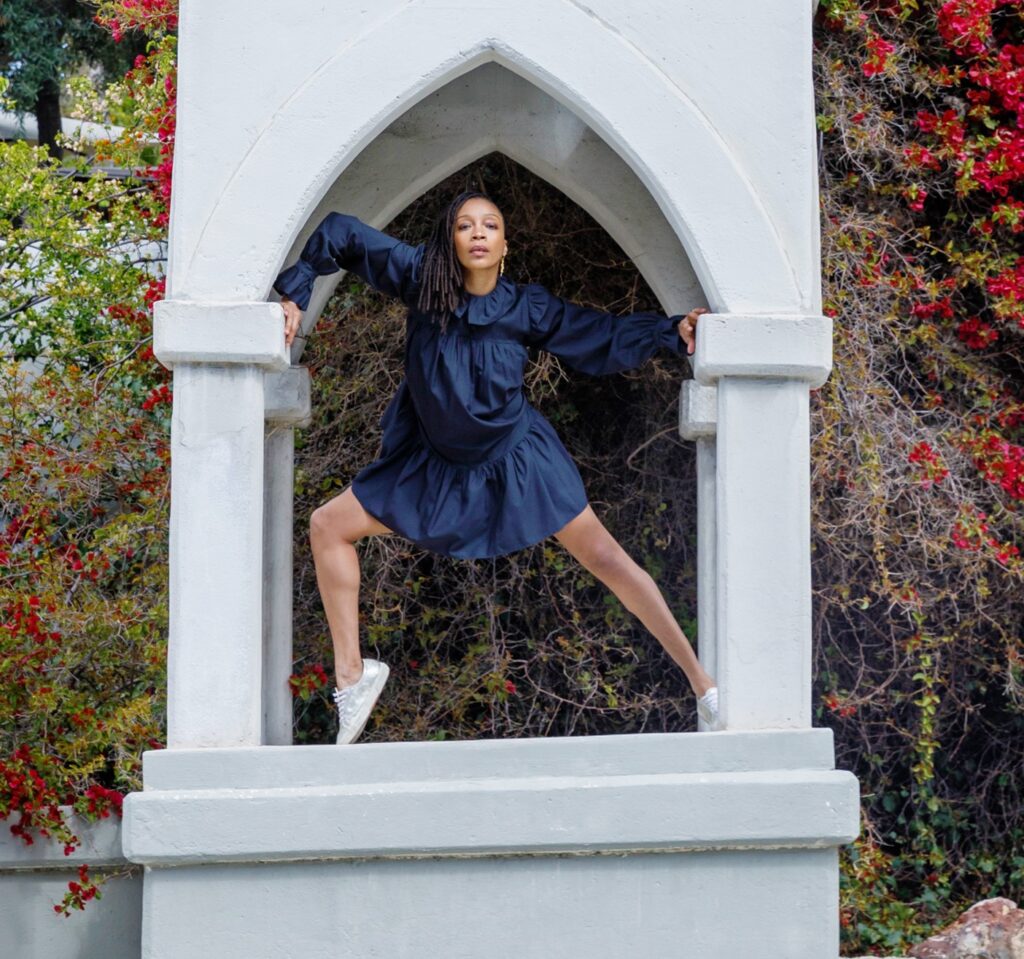 a female dancer wearing a long sleeved blue dress and white sneakers posing underneath an archway