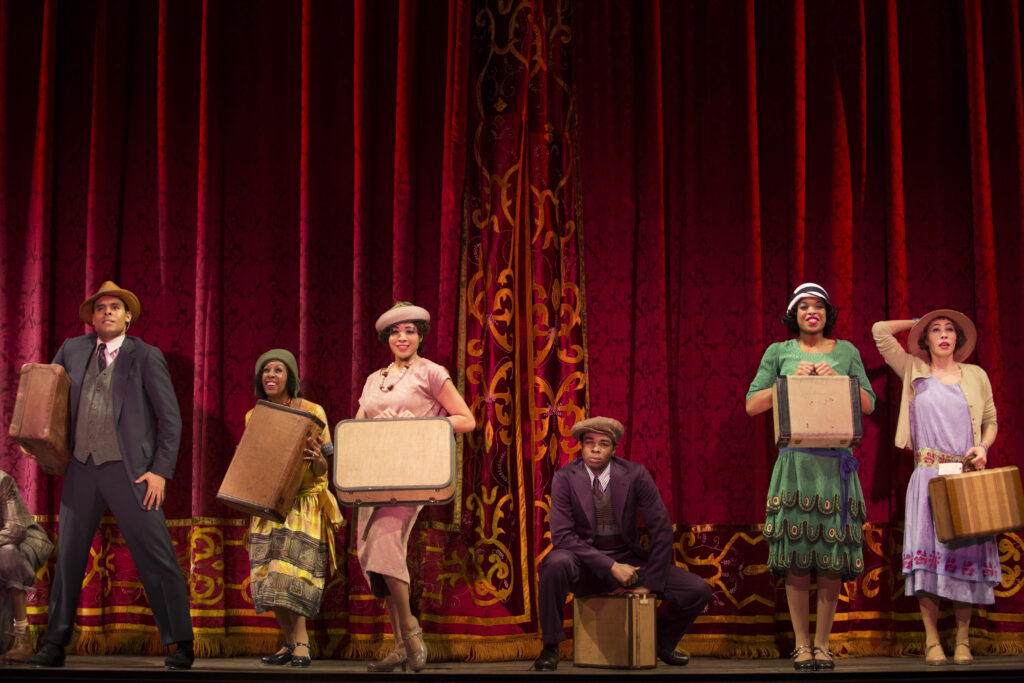 Karissa Royster stands with a beveled foot, an old fashioned suitcase raised in front of her stomach. She wears an old fashioned pale pink dress, a matching hat, and heeled tap shoes. She poses in front of a red curtain with gold filigree, surrounded by other performers in similar period dress.