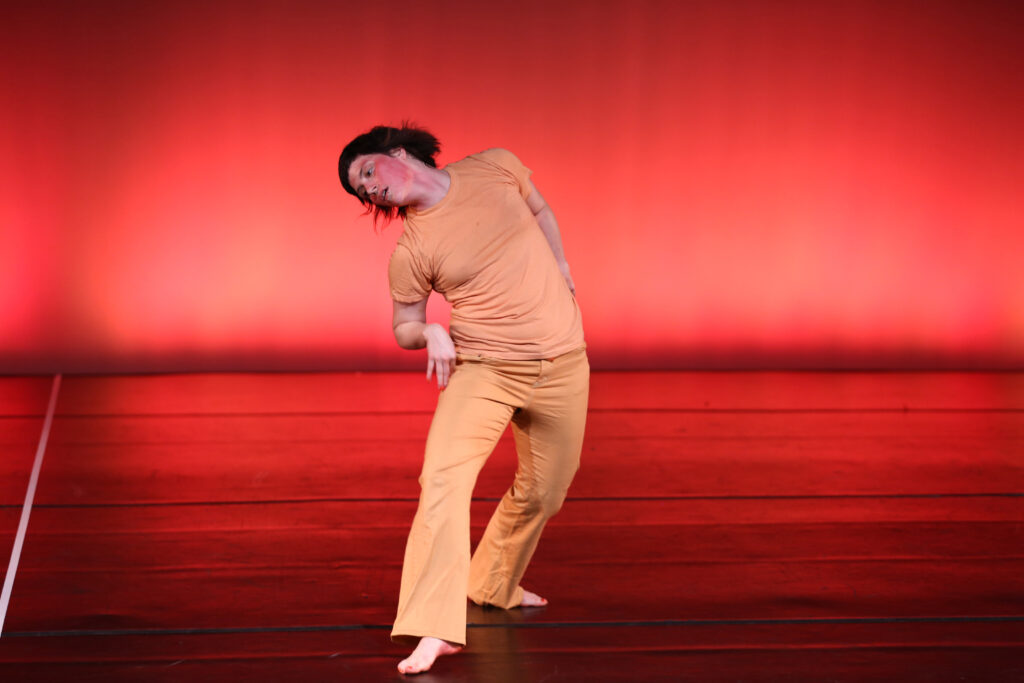 Meg Foley is caught mid-performance. She faces forward in a wide fourth position plié, leaning to her right. She wears pale orange pants and a shirt, while the stage is lit in red.