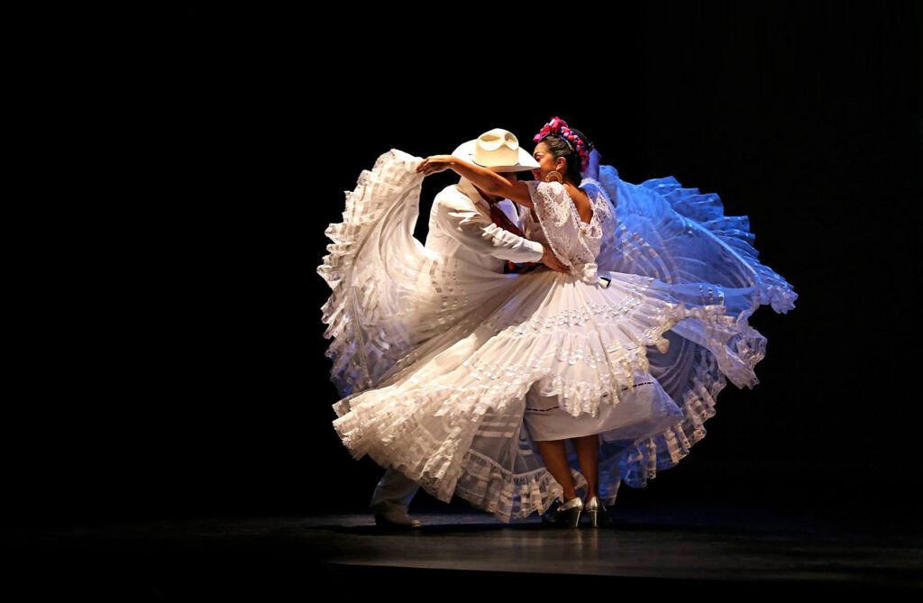 A diaphanous white dress flares up and around as the woman wearing it is whirled around by a man in a white suit.