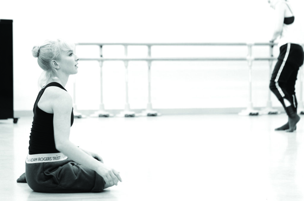 A black and white image of Charlotte Edmonds sitting on the floor of a studio. Her blonde hair is pulled back into a bun. She is shown in profile, posture straight as she watches intently.