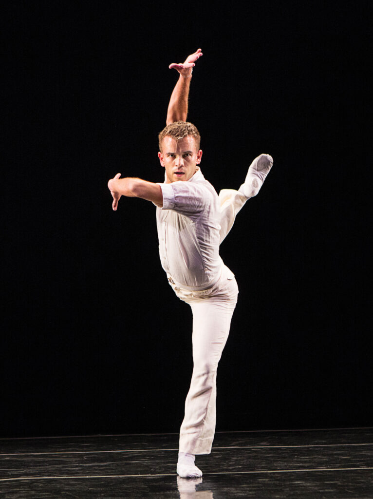 Reed Tankersley directs an intense look outward as he moves through a back attitude in plié. His arms curve and wrap around his torso, palms turned outward. He wears a white short sleeved shirt, white pants, and white ballet slippers.